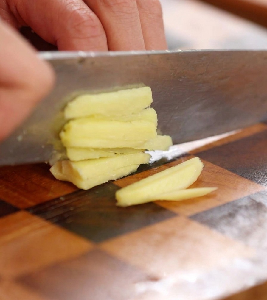 Primer plano de un cuchillo picando finamente una rodaja de jengibre fresco en trozos diminutos sobre una tabla de madera.