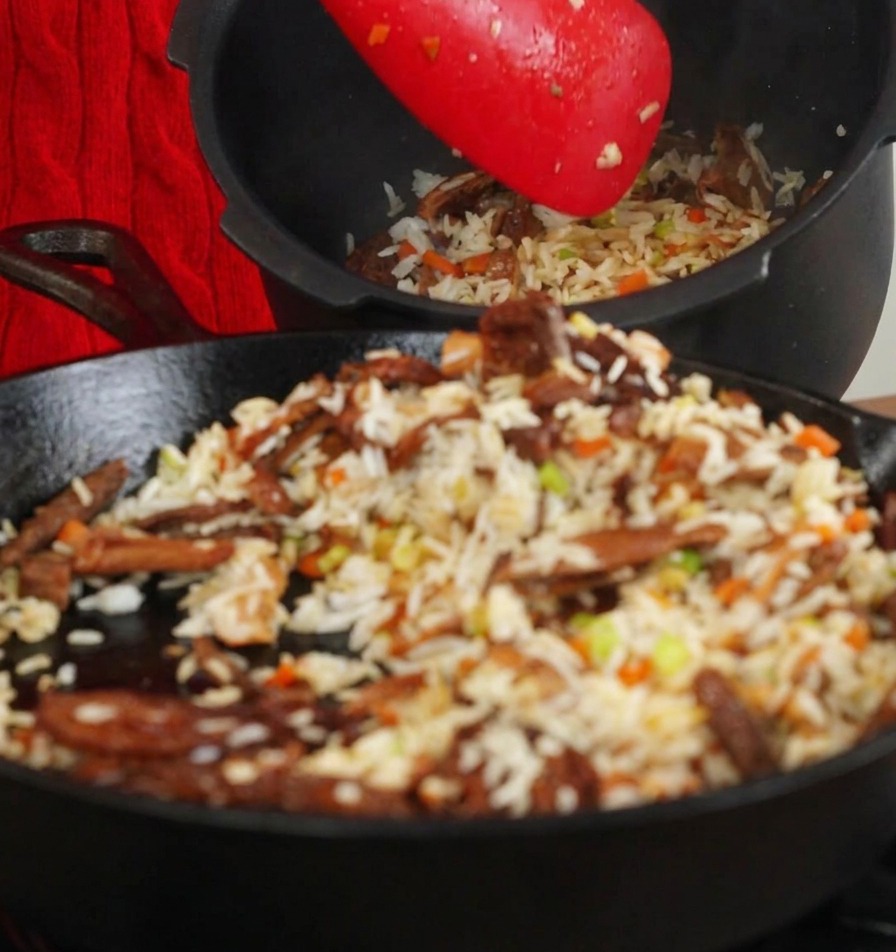 Uso de una espátula roja para pasar el arroz salteado con setas desde la sartén al recipiente interno de una arrocera negra.
