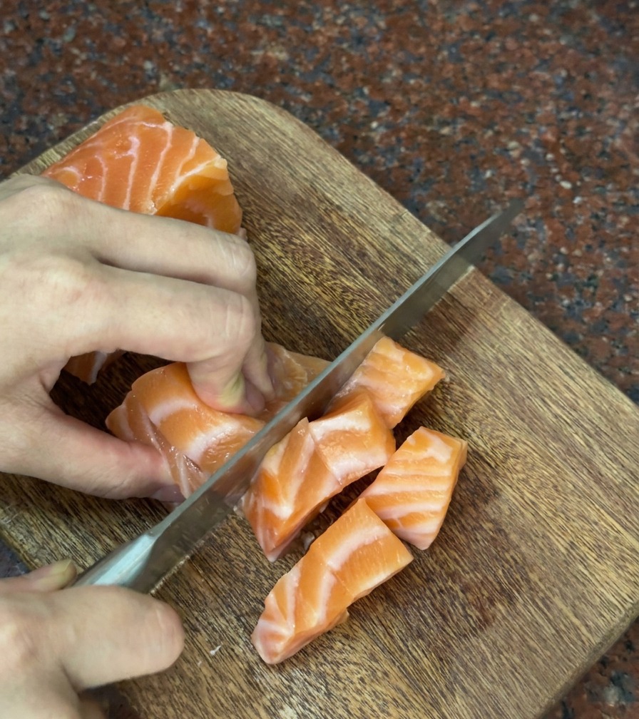 Manos cortando cuidadosamente un filete de salmón naranja en cubos uniformes sobre una tabla de madera.