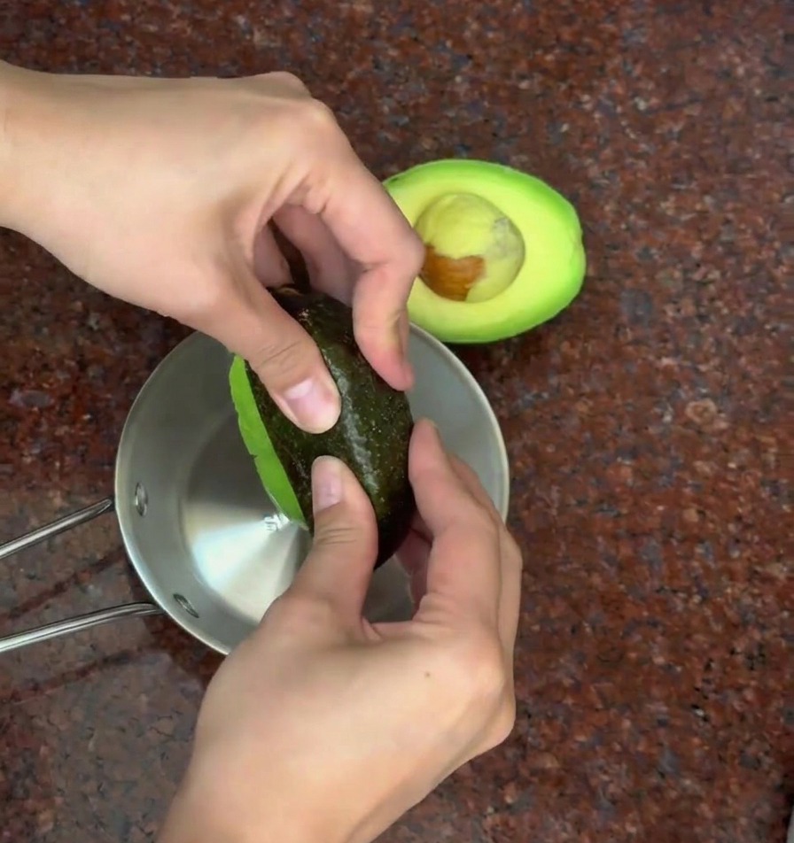 Persona retirando la piel oscura de la pulpa verde de un aguacate a la mitad sobre un bol de metal.
