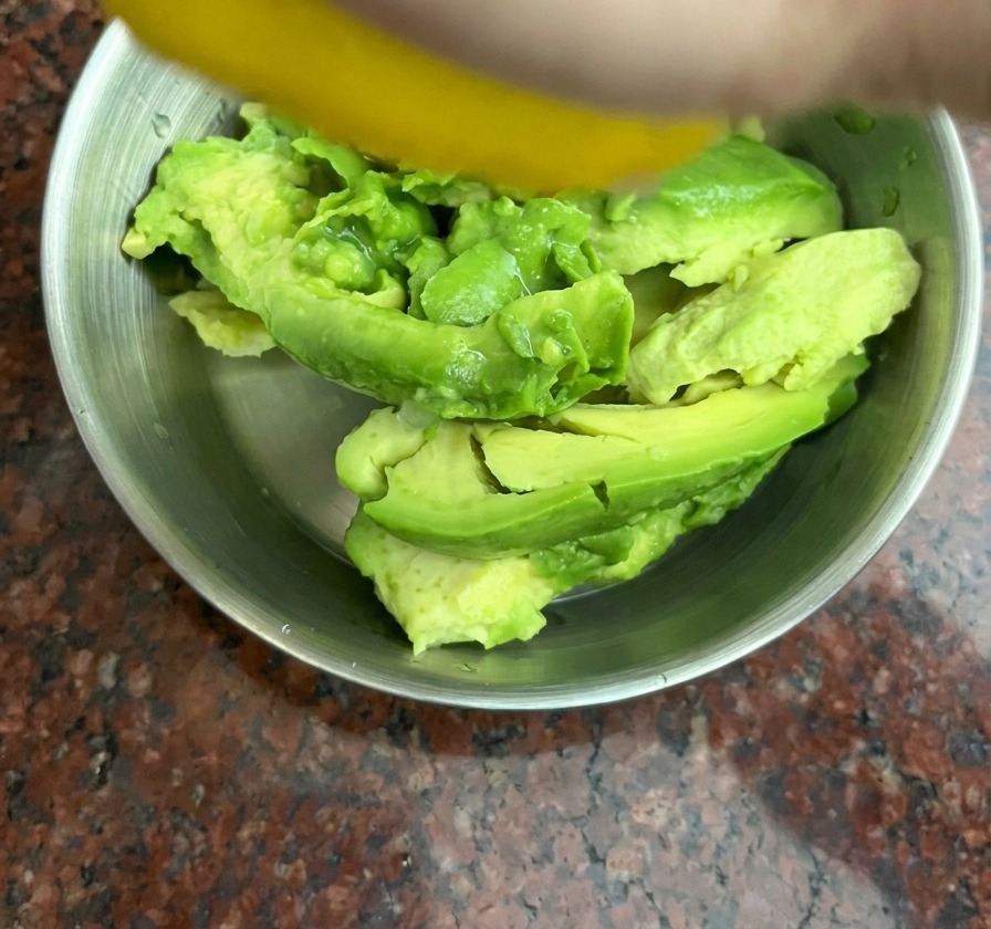Mano exprimiendo un limón amarillo sobre trozos de aguacate en un bol de acero inoxidable.