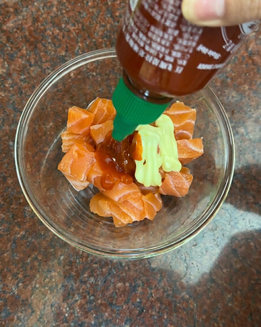 Añadiendo mayonesa y salsa de chile rojo sobre los cubos de salmón crudo.