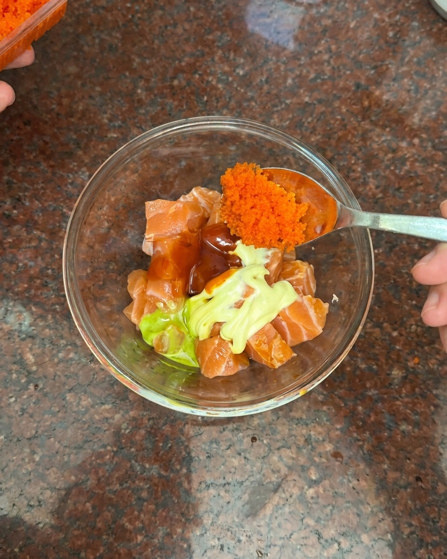 Cuchara añadiendo una porción de huevas de pescado naranjas a un bol con salmón, mayonesa y salsa de chile.