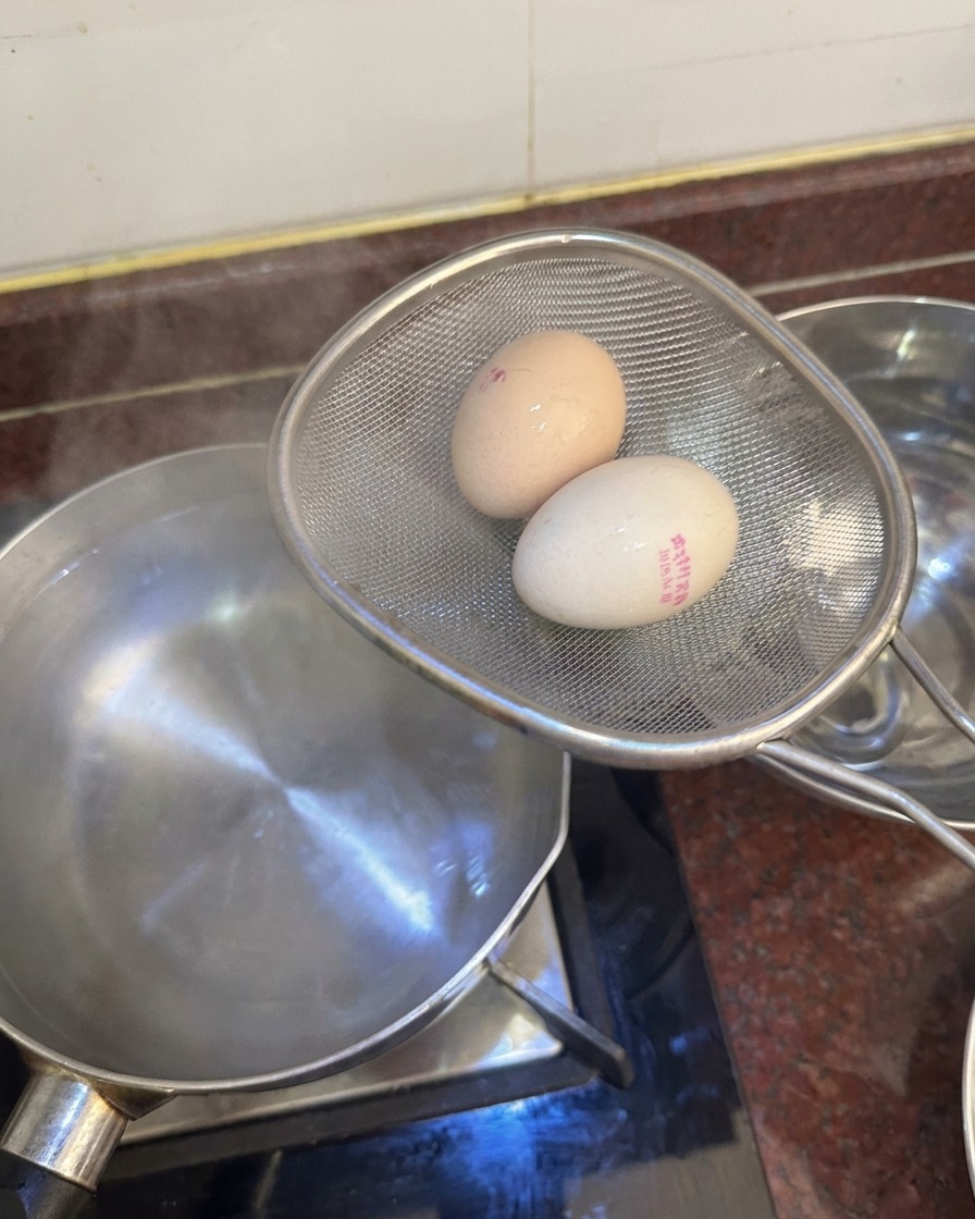 Colador metálico con dos huevos sobre una olla, listos para pasar a un bol cercano con agua y hielo.