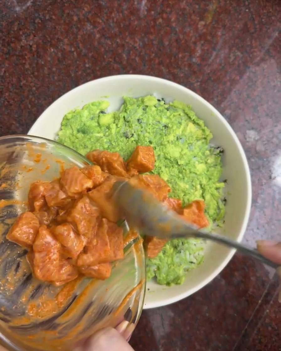 Cubos de salmón marinado pasando de un bol de cristal a la capa de aguacate en un bol blanco.