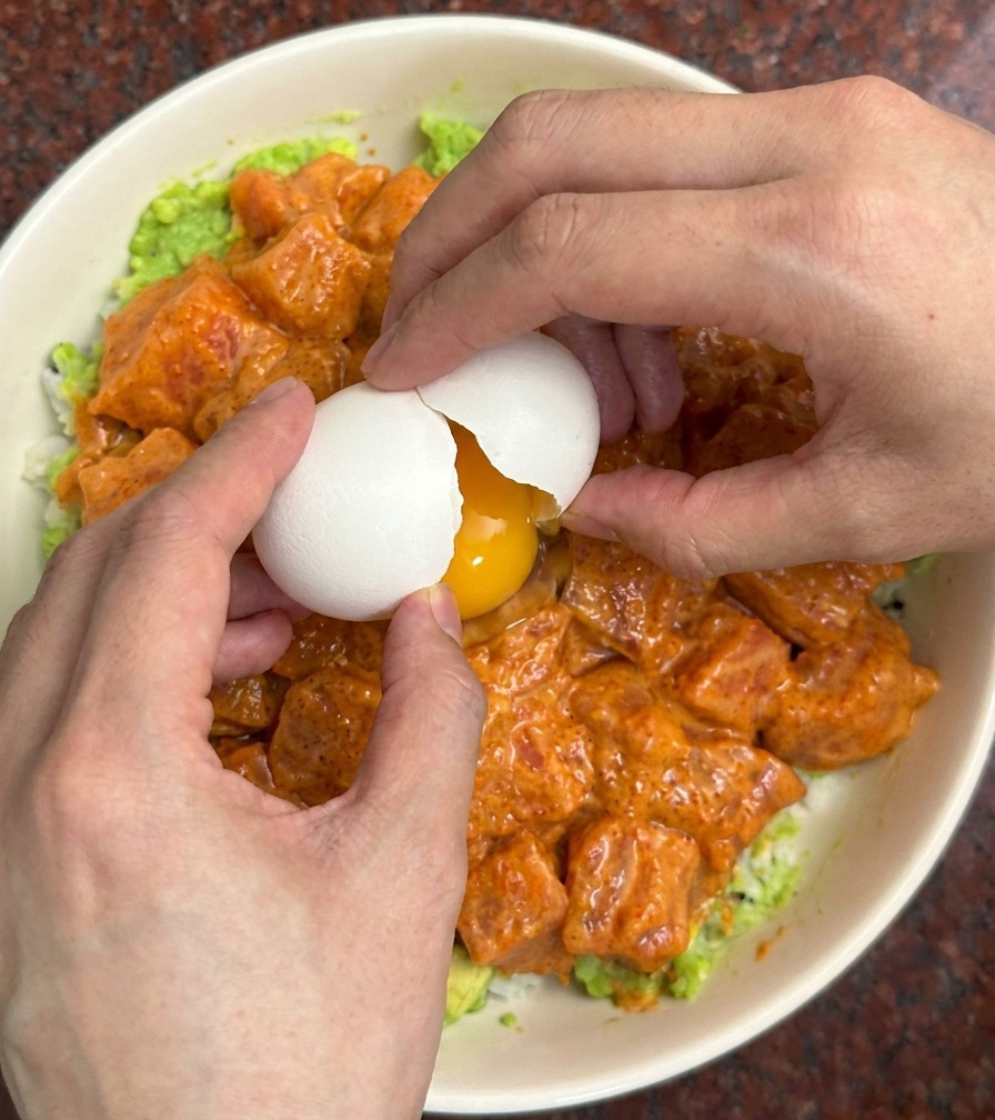 Manos rompiendo un huevo blanco sobre un bol colorido lleno de salmón marinado y aguacate.