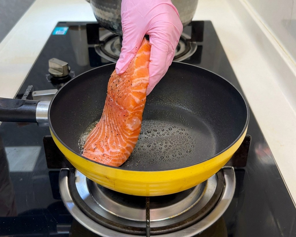 Un filete de salmón crudo sazonado siendo colocado en una sartén amarilla caliente por una mano con guante rosa.