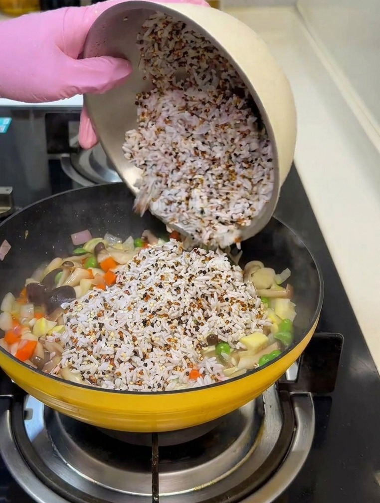 Una mano con guante rosa vertiendo una mezcla de arroz blanco y quinoa en la sartén con los vegetales salteados.