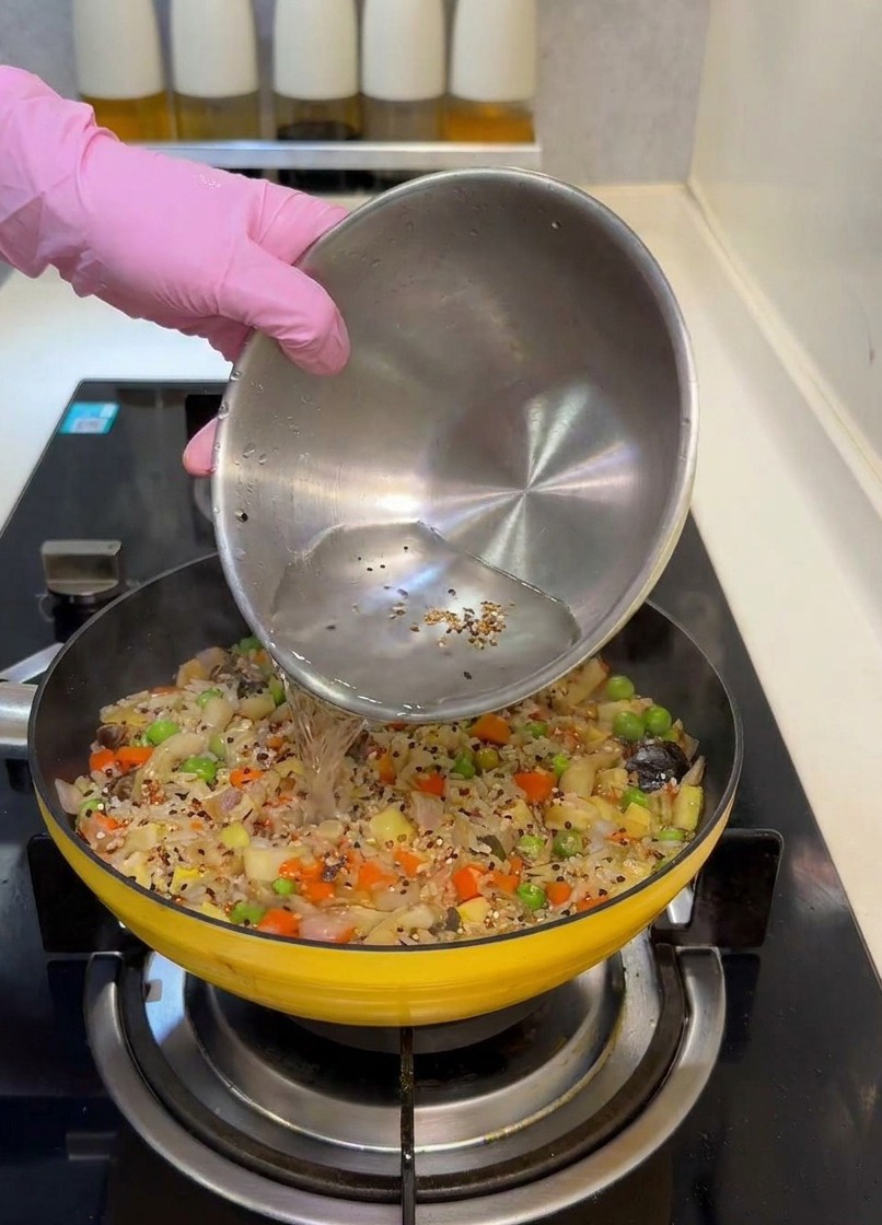 Agua vertiéndose desde un bol de acero inoxidable a la sartén llena de arroz y vegetales.