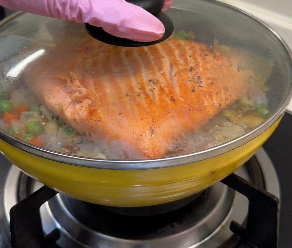 Una sartén amarilla con arroz y salmón siendo tapada con una tapa de cristal para atrapar el vapor.