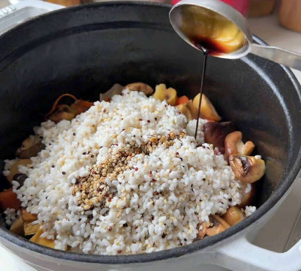 Cuchara metálica vertiendo salsa de soja sobre una montaña de arroz y quinoa con setas en una olla negra.