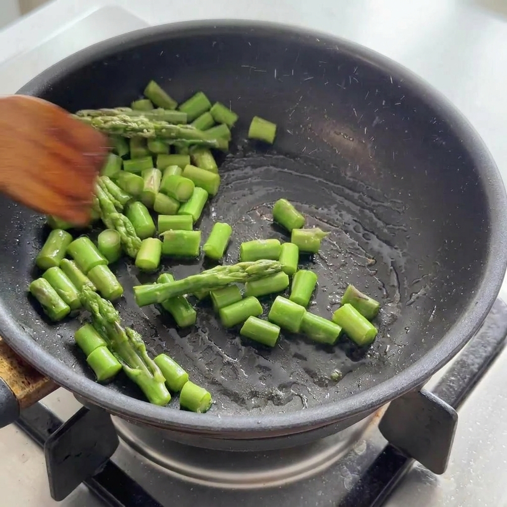 Trozos de espárragos verdes salteándose en una sartén negra con una espátula de madera.