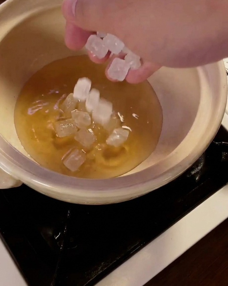Una mano dejando caer cristales de azúcar cande translúcidos en una olla con mirin.