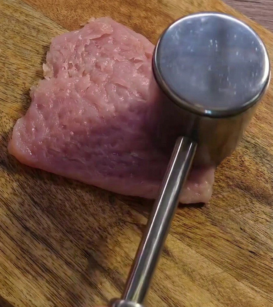 Un mazo metálico para carne golpeando una rodaja de cerdo crudo en una tabla de madera para aplanarla.