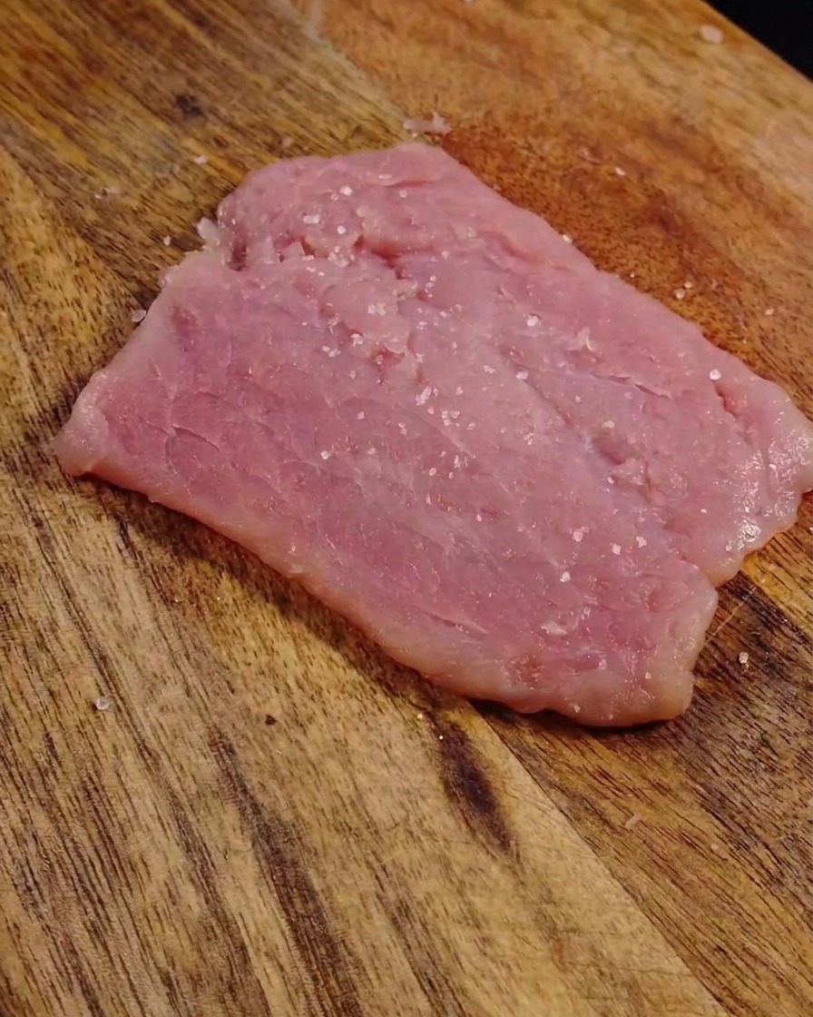 Granos de sal y pimienta negra visibles sobre la superficie de una rodaja de cerdo cruda, aplanada y tierna.