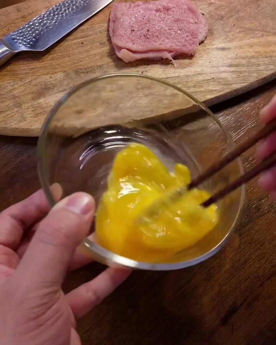 Una persona usando palillos de color marrón oscuro para batir huevos crudos en un bol de cristal sobre una mesa de madera.