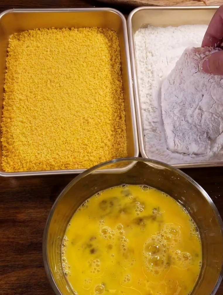 Una mano pasando una chuleta de cerdo sazonada por una bandeja rectangular con polvo de maicena blanca.
