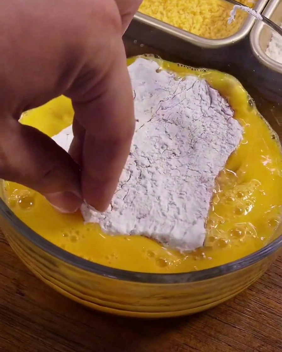 Una pieza de cerdo cubierta de maicena blanca siendo sumergida en un bol de huevos batidos amarillos.