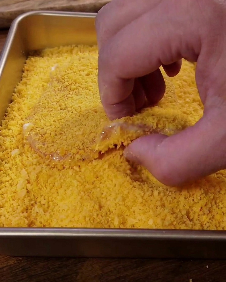 Presionando una pieza de cerdo en una bandeja llena de pan rallado Panko de color amarillo dorado.