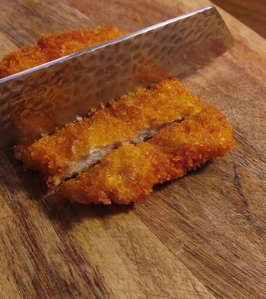 Primer plano de una hoja de cuchillo metálica cortando una chuleta de cerdo frita y crujiente sobre una tabla de madera.