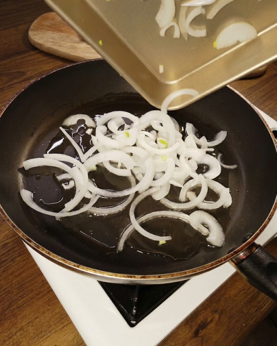 Aros de cebolla blanca cortados vertiéndose desde una bandeja dorada en una sartén antiadherente con aceite brillante.