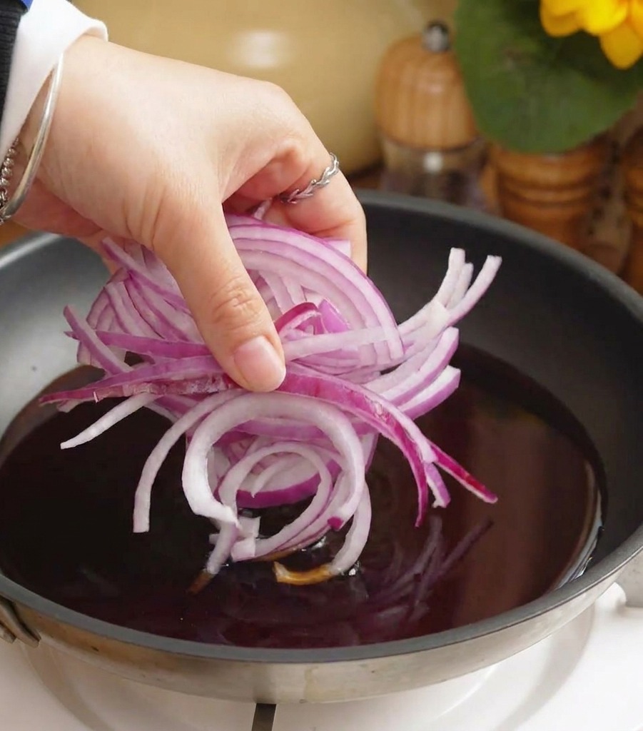 Una mano echando cebolla morada laminada en una sartén con salsa oscura hirviendo.
