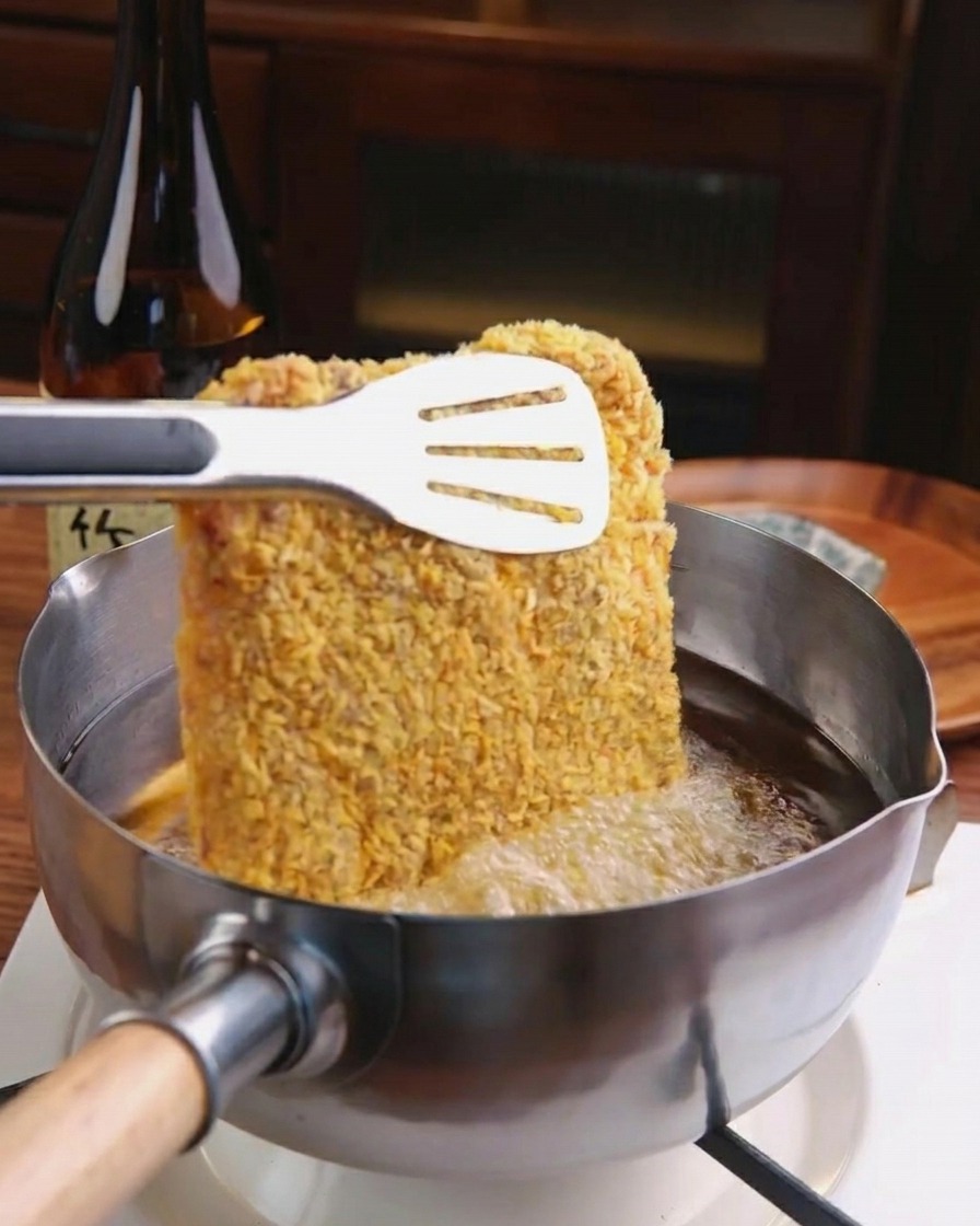 Una chuleta de cerdo empanizada siendo introducida en una olla con aceite caliente burbujeante usando pinzas de metal.
