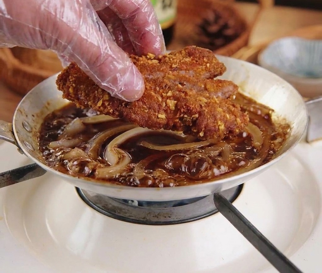 Una mano con guante colocando una chuleta de cerdo frita y cortada en una sartén con cebollas y salsa hirviendo.