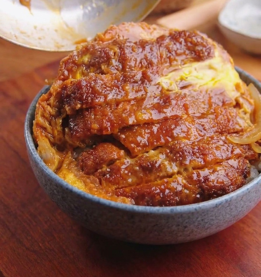 Una sartén inclinándose sobre un bol de arroz para deslizar una chuleta de cerdo con huevos y cebollas.