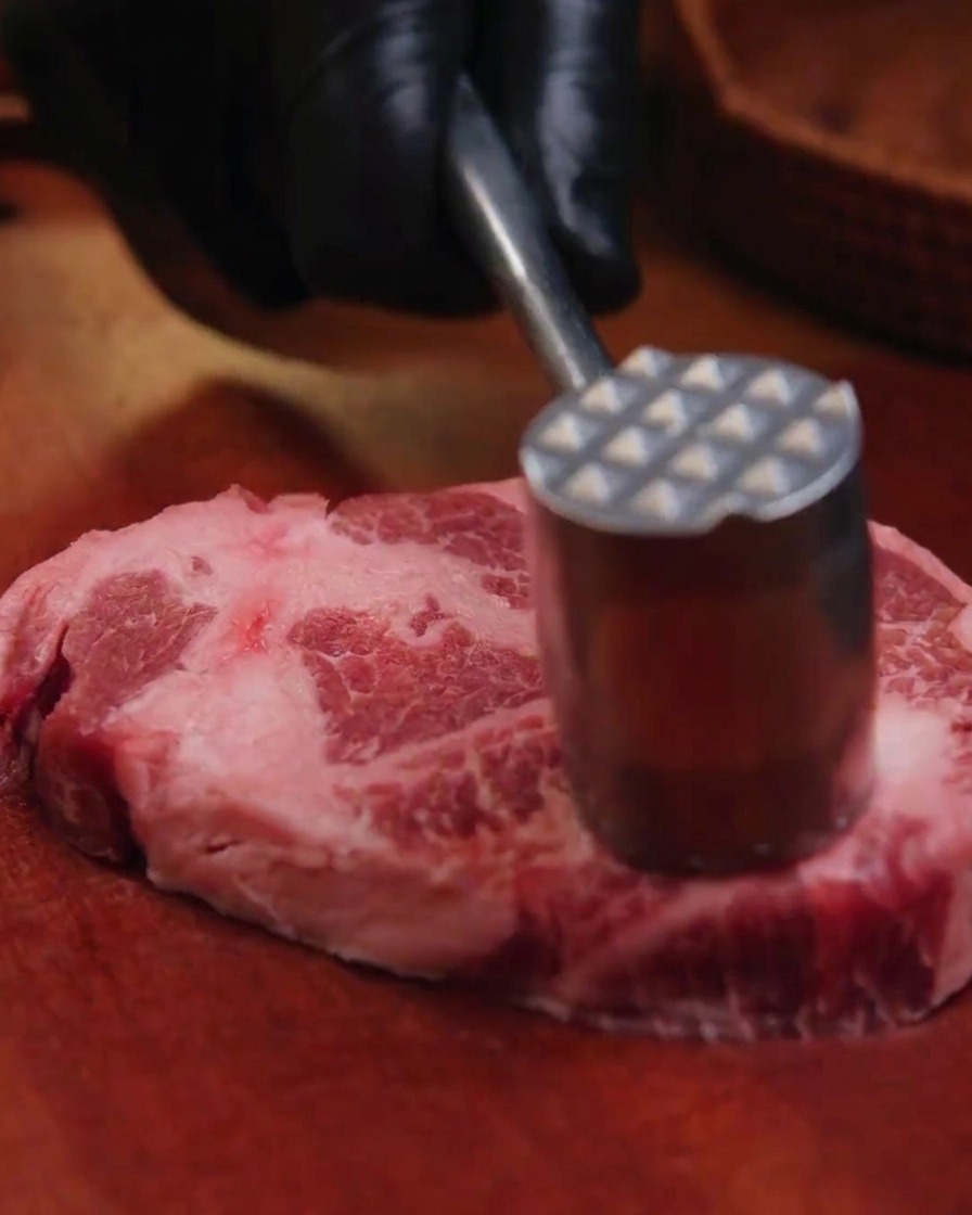 Mano con guante negro golpeando una pieza de aguja de cerdo con un mazo de carne sobre una tabla de madera.
