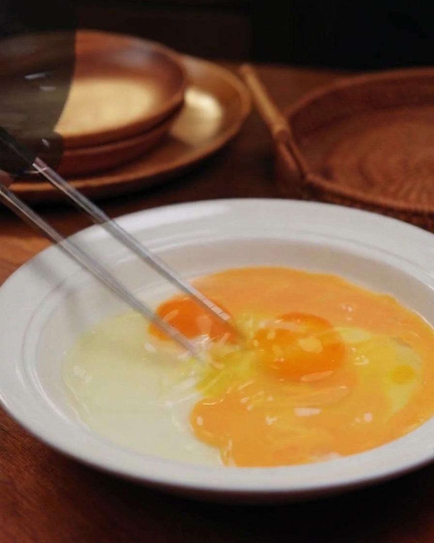 Palillos batiendo dos huevos en un cuenco blanco, creando una mezcla veteada de yema y clara.
