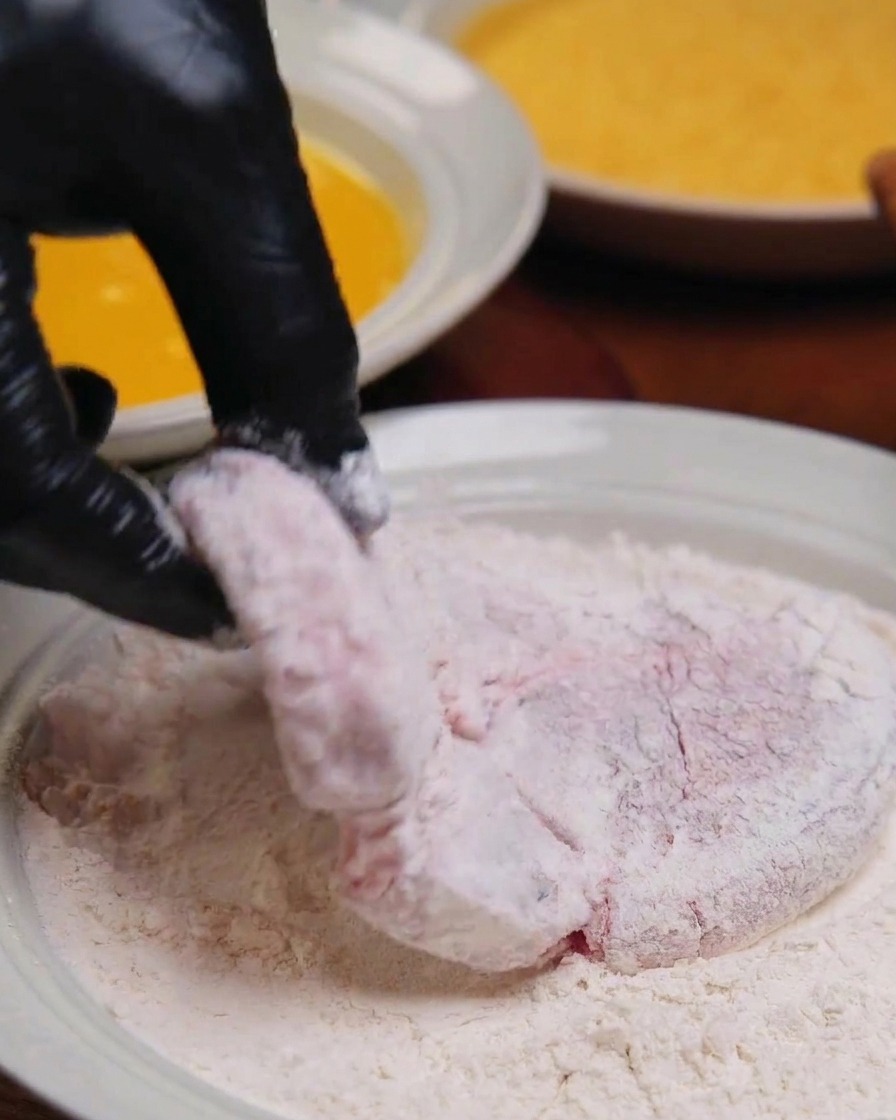 Mano enguantada sosteniendo una pieza de cerdo totalmente cubierta de harina blanca sobre un plato.