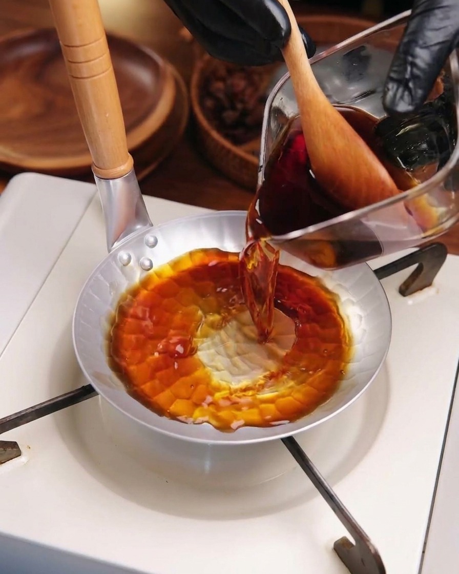 Salsa oscura para katsudon vertiéndose desde una jarra a una pequeña sartén metálica al fuego.