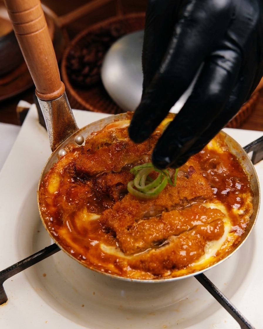 Mano colocando aros de cebolleta fresca sobre la mezcla de Katsudon cocinada en la sartén.