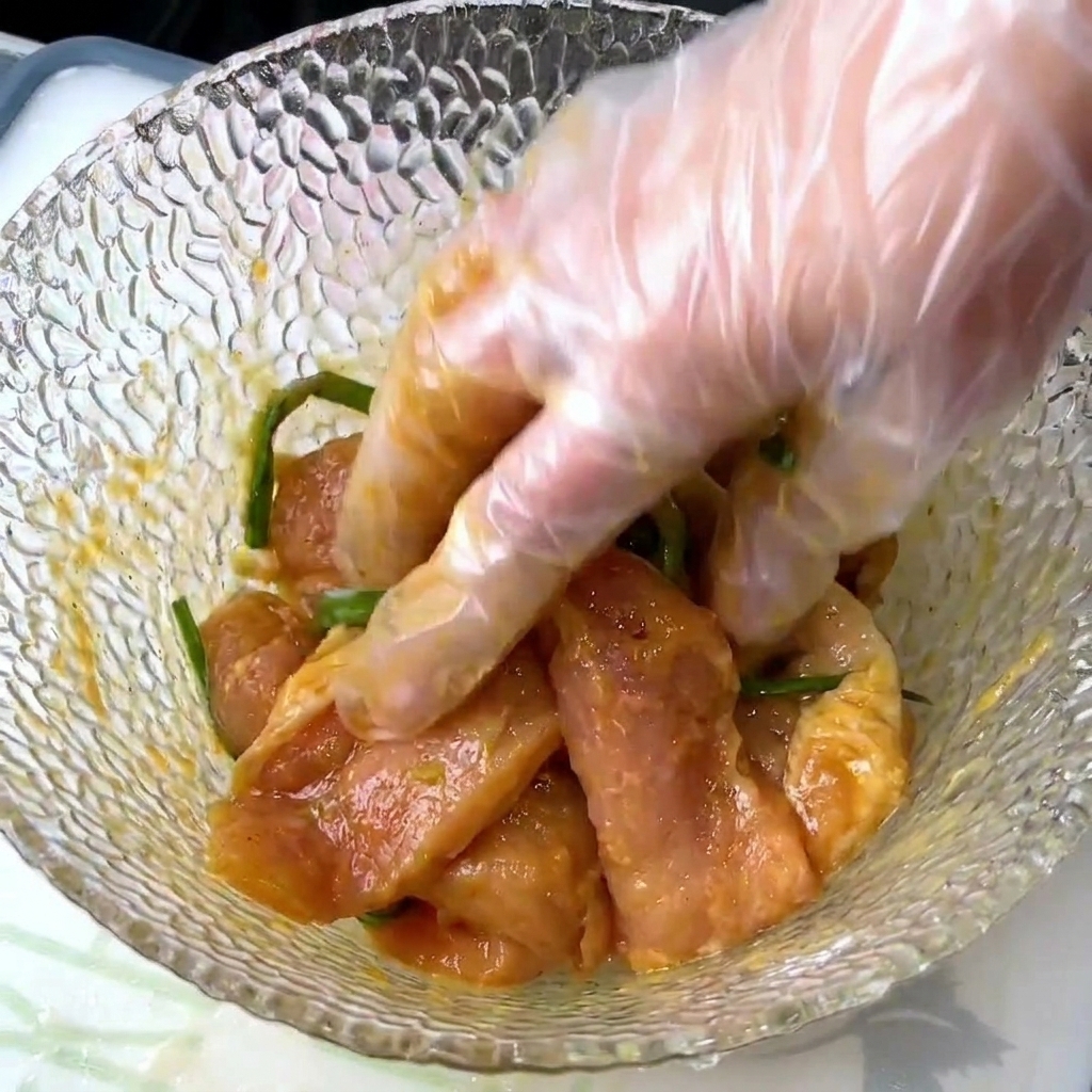 Mano con guante mezclando rodajas de cerdo con cebolletas y condimentos líquidos en un bol de cristal tallado