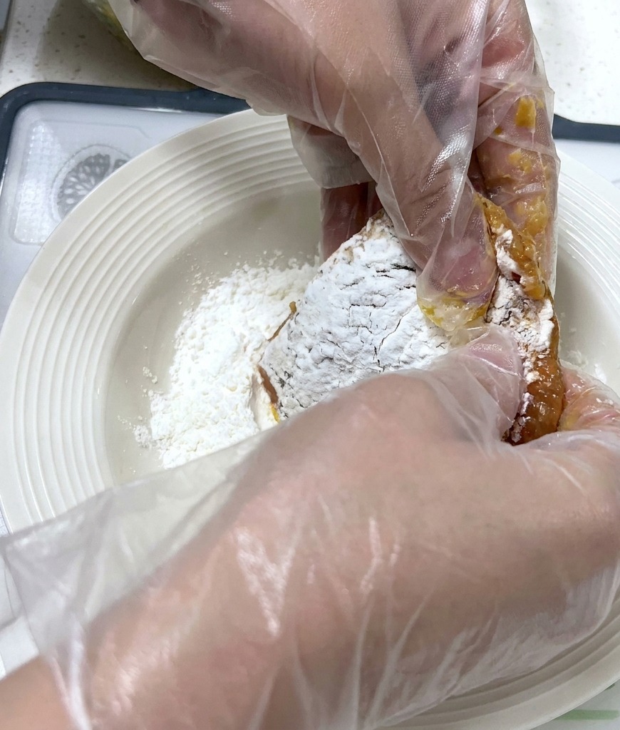 Manos con guantes presionando una chuleta de cerdo marinada en un plato con almidón blanco en polvo
