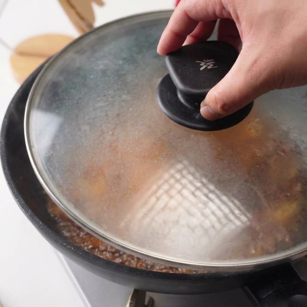 Una mano colocando una tapa de cristal transparente en una sartén con pollo y salsa hirviendo.