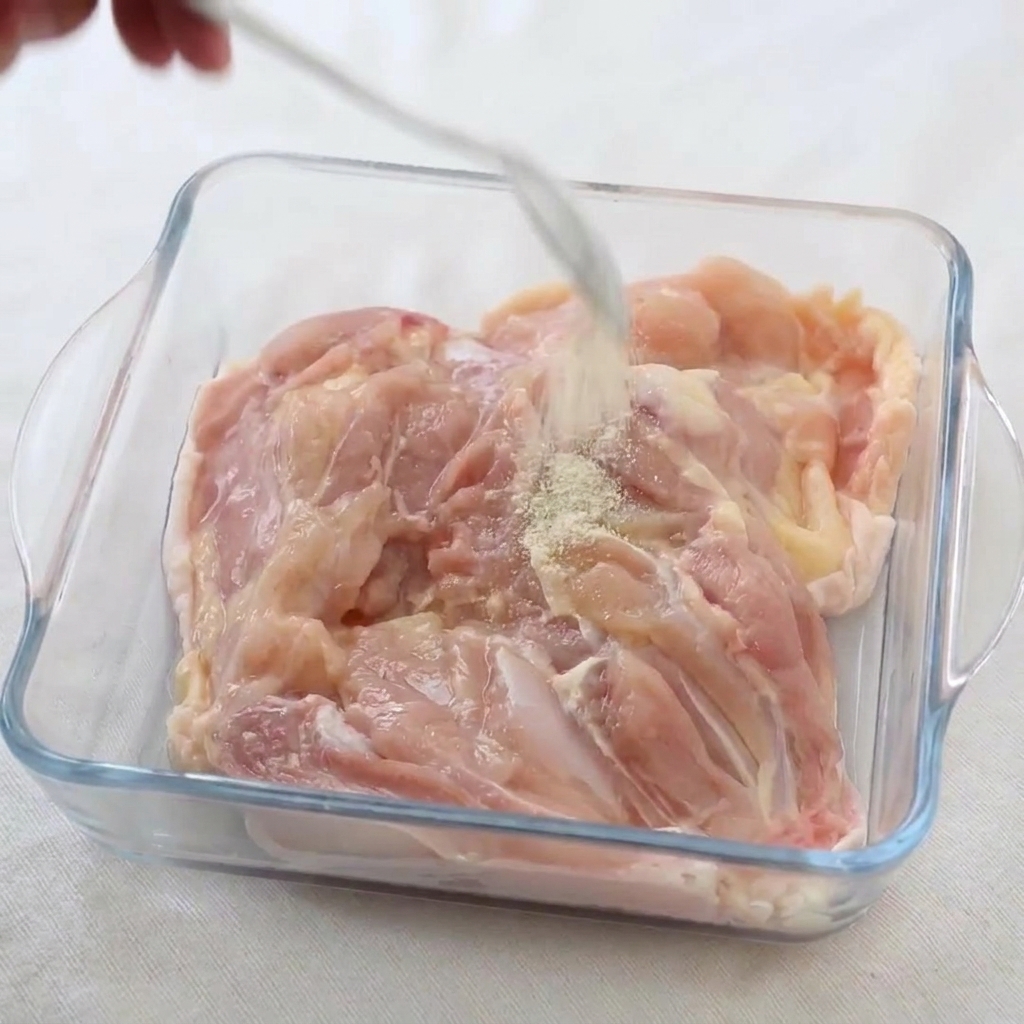 Pimienta blanca y sal siendo espolvoreadas desde una cuchara sobre contramuslos de pollo en un recipiente de vidrio.