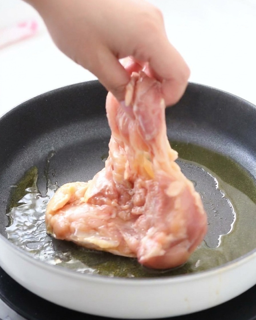 Una mano colocando un contramuslo marinado con la piel hacia abajo en una sartén caliente con aceite brillante.