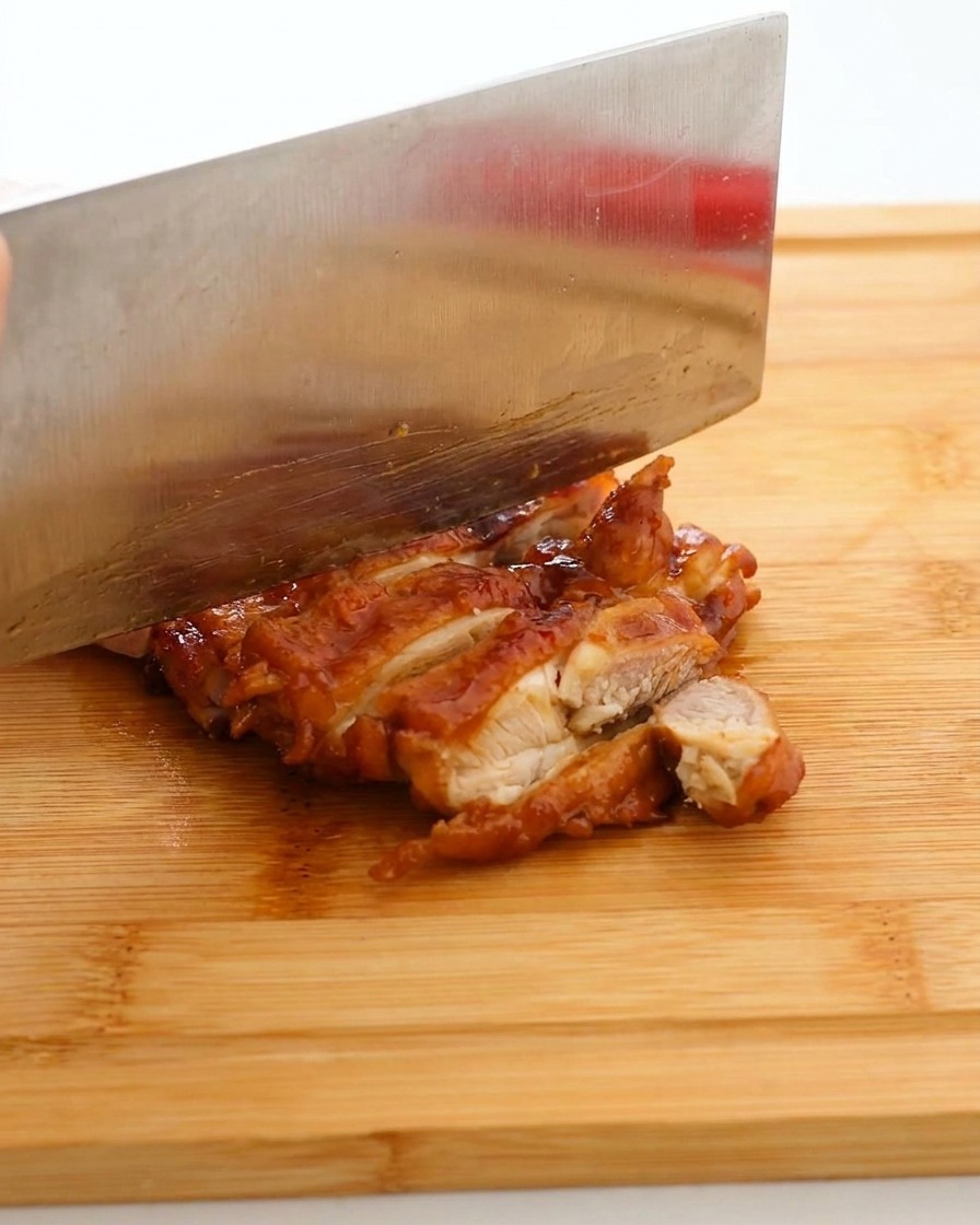 Un cuchillo de cocina grande cortando un contramuslo glaseado en tiras sobre una tabla de madera.