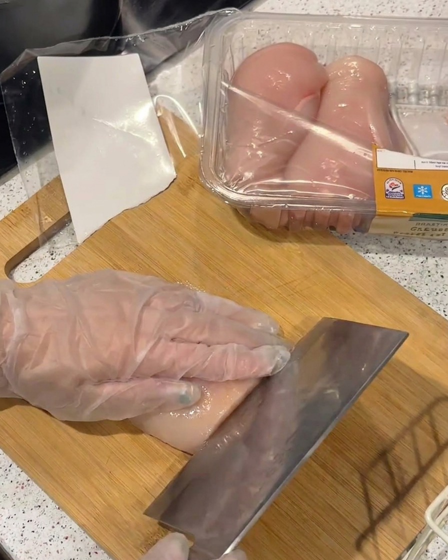 Una mano con guante de plástico cortando una pechuga de pollo cruda en filetes finos con un cuchillo de cocina sobre una tabla de madera.