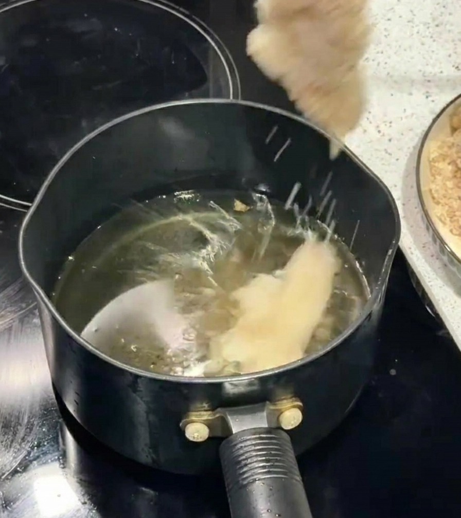 Una mano usando palillos para introducir un filete de pollo empanado en una olla con aceite hirviendo.