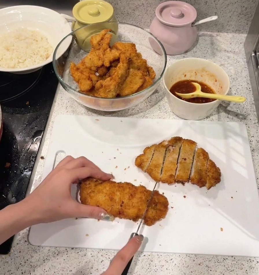 Una persona cortando un filete de pollo frito dorado en tiras sobre una tabla de cortar blanca.