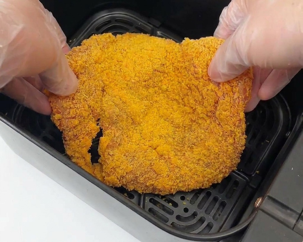 Dos manos con guantes colocando una pieza grande de pollo empanizado en la canasta negra de una freidora de aire.