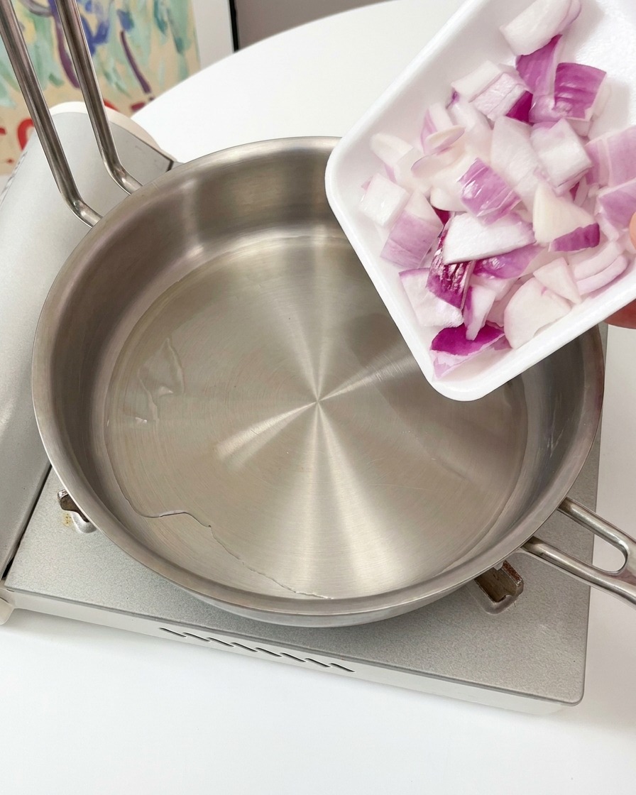 Una mano vertiendo cebollas moradas picadas desde una pequeña bandeja cuadrada en una sartén de acero inoxidable con aceite caliente en la estufa.