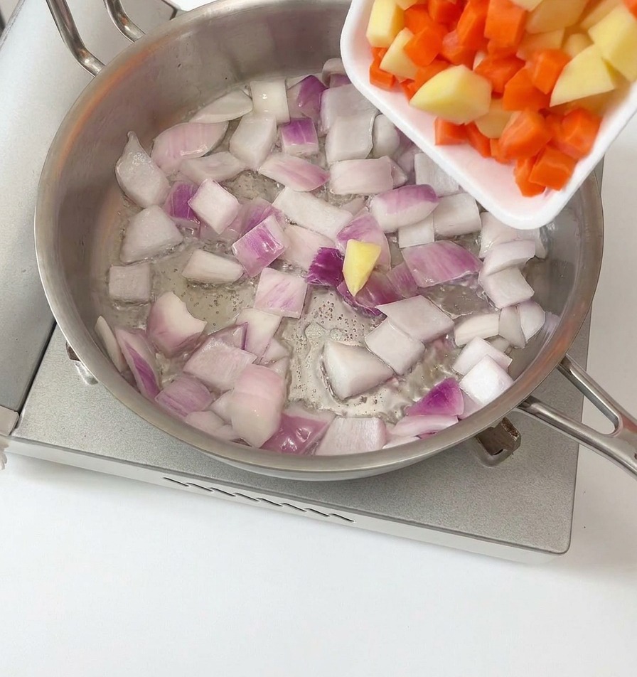 Papas y zanahorias en cubitos siendo vertidas desde una bandeja blanca a una sartén de acero inoxidable que contiene cebollas moradas sofritas.