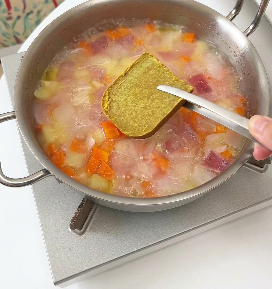 Pinzas de metal sosteniendo un cubo cuadrado y sólido de curry marrón sobre una olla humeante con vegetales y agua.