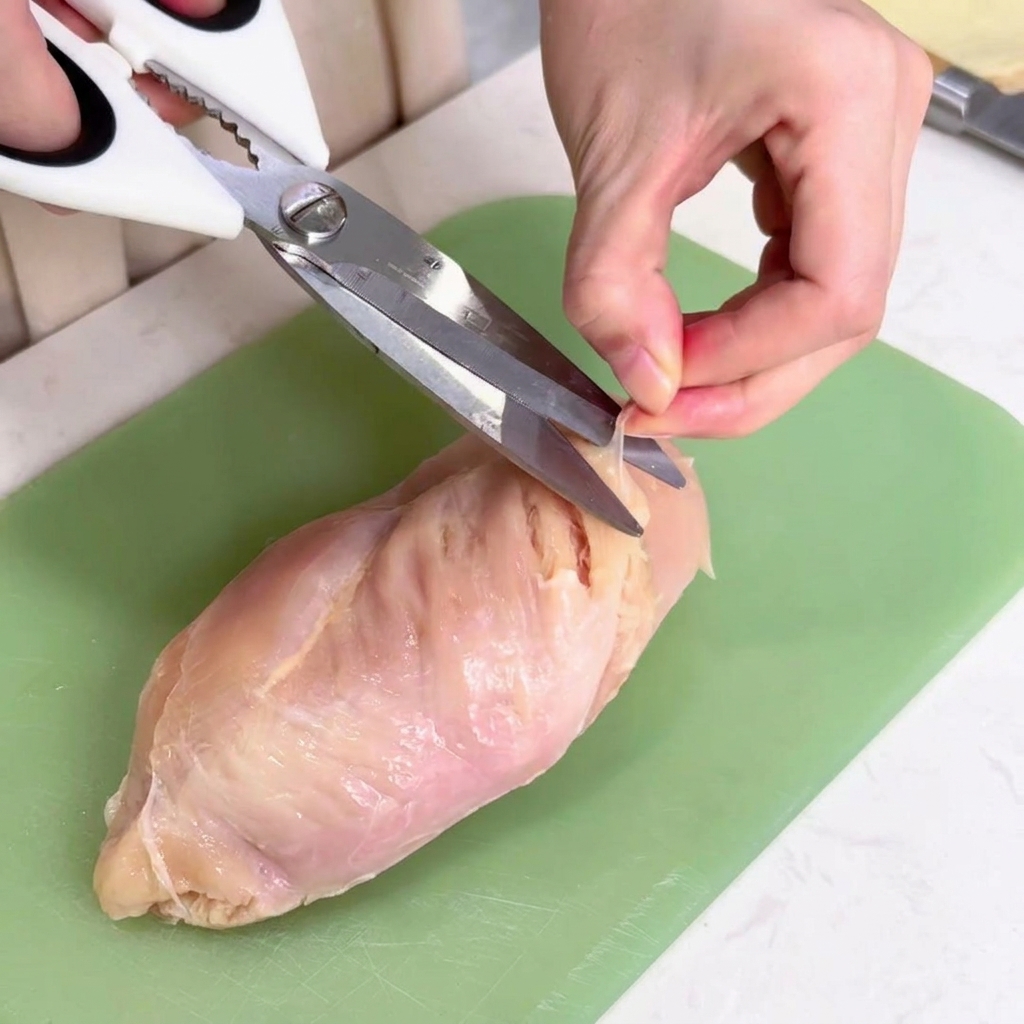 Uso de tijeras de cocina blancas para quitar el tejido conectivo transparente de una pechuga de pollo sobre una tabla de cortar verde.