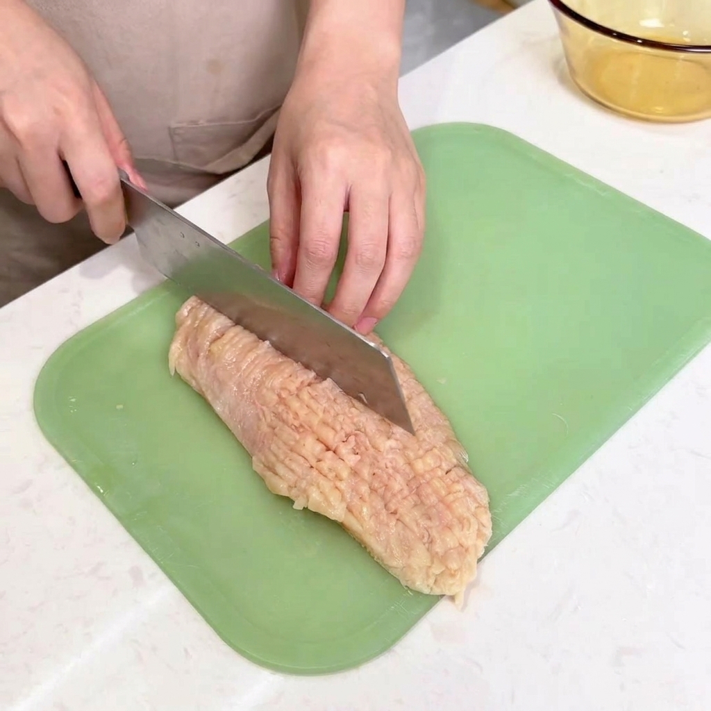 El lomo de un cuchillo de chef golpeando una pechuga de pollo abierta para ablandarla sobre la tabla de cortar.