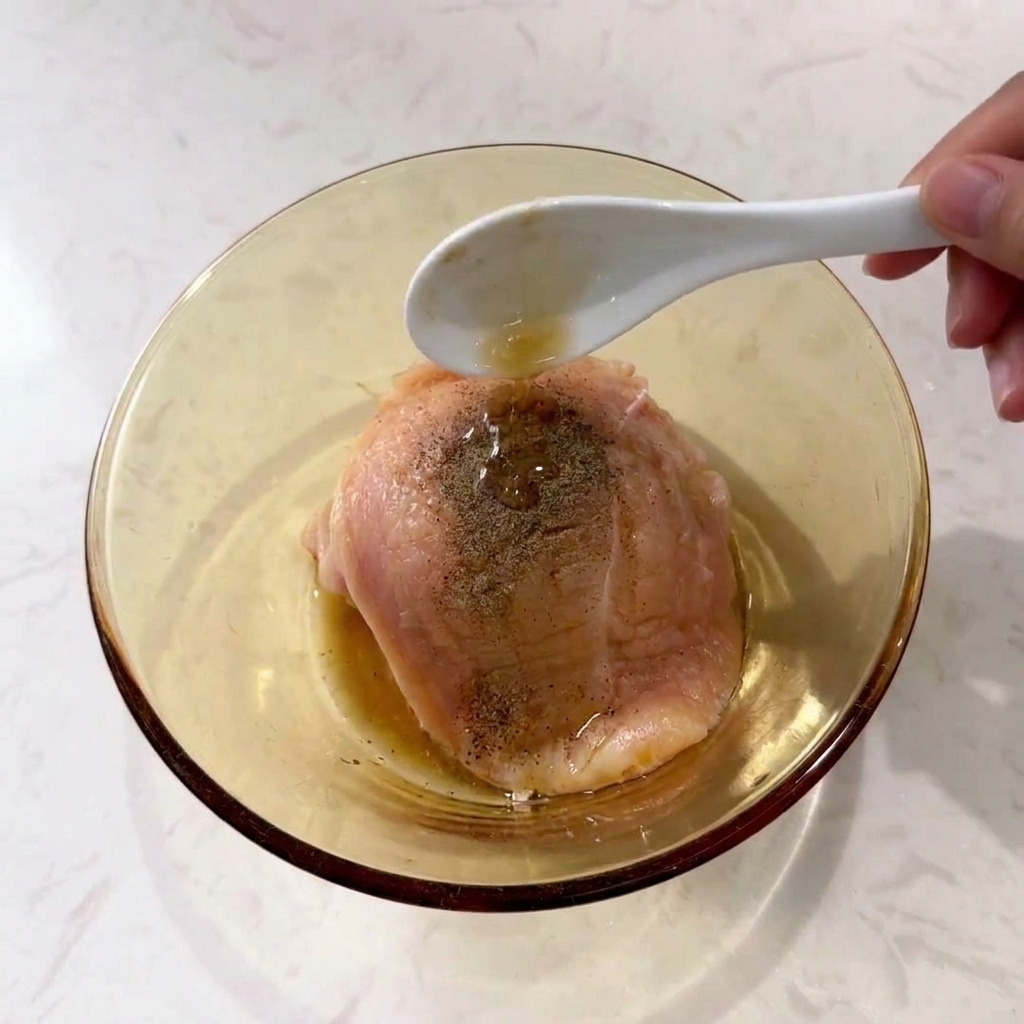 Cuchara vertiendo condimento líquido sobre pechuga de pollo con pimienta negra en un bol de cristal.